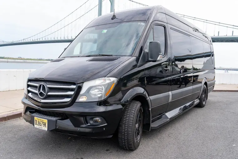 A black Mercedes-Benz Sprinter executive van parked by the water beneath a suspension bridge