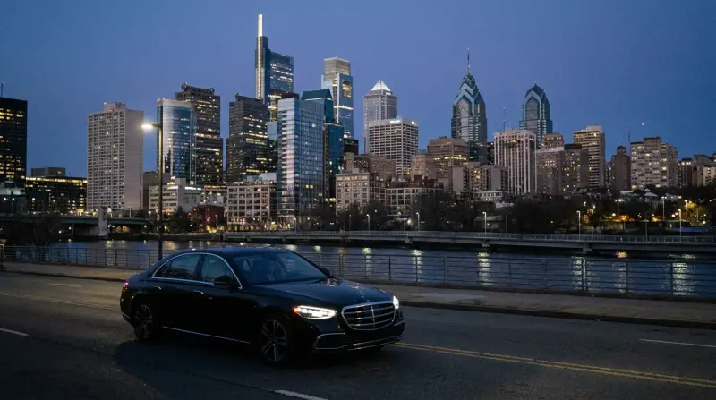 A black sedan on the riverfront with the Philadelphia skyline lit at dusk