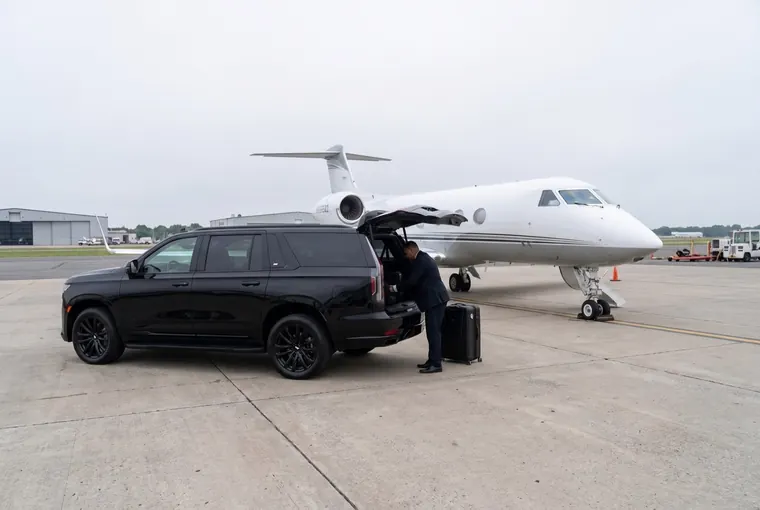 A chauffeur loading luggage into a black Cadillac Escalade parked beside a private jet on the tarmac